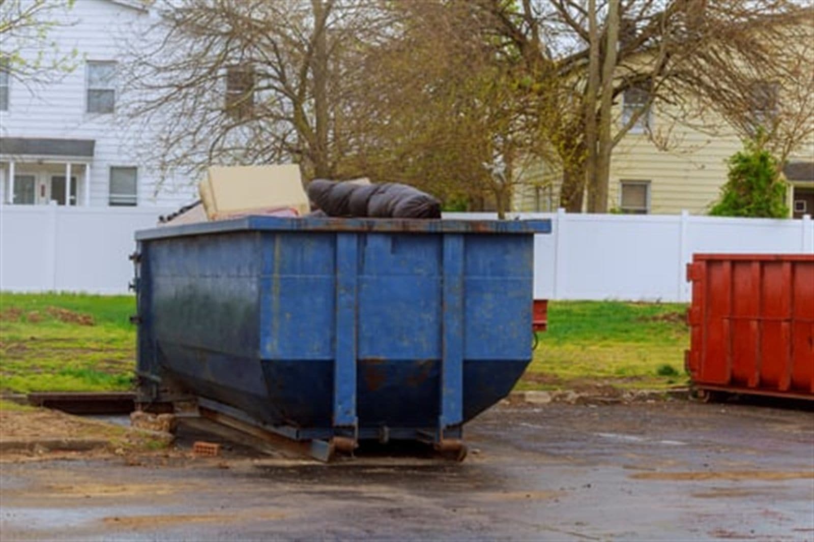 A blue dumpster loaded with discarded furniture and moving debris outside a residential home